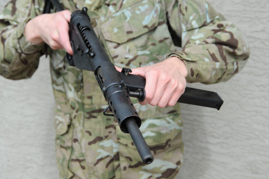 A soldier, whose face is not seen, holds a sten gun.