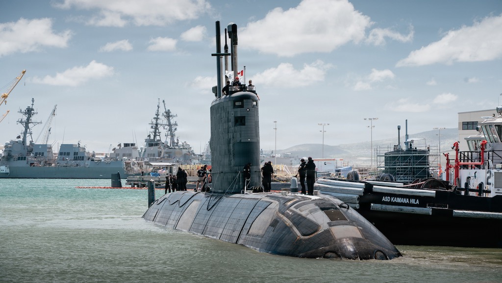 A submarine is partly submerged in a busy port. Several people work on its outer surface.