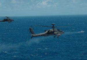 Two helicopters fly above the sea toward a ship in the distance.