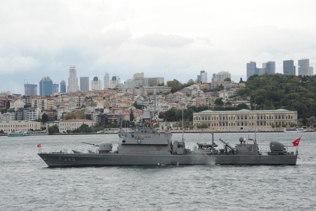 A military ship is seen in the water with a cityscape in the background.